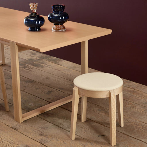 Runder Hocker Punton aus hellem Massivholz mit gepolsterter Sitzfläche neben Holztisch auf rustikalem Holzboden im Wohnbereich