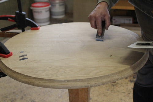 Ein Handwerker spachtelt eine runde Tischplatte aus Massivholz in sorgfältiger Handarbeit. Diese Bearbeitung sorgt für eine glatte Oberfläche und eine hochwertige Basis für langlebige Beizentische, Bistrotische oder Restauranttische nach Maß.